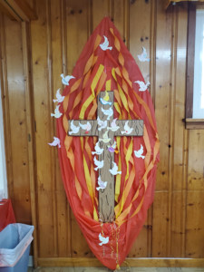 Lifesized flame made with red, orange and yellow craft paper.  A cardboard cutout of a cross lies in the center while small dove cutouts are sprinkled around on a wall of the parish house.